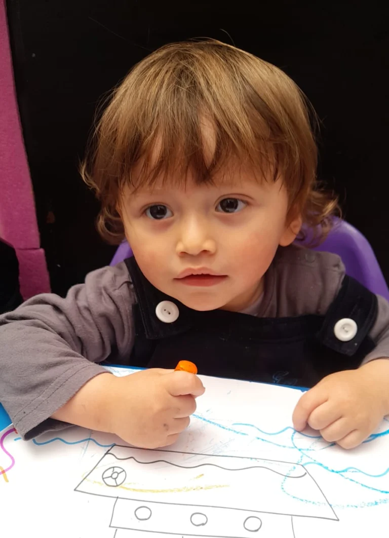 Niño pequeño dibujando en una hoja de papel con un crayón naranja.