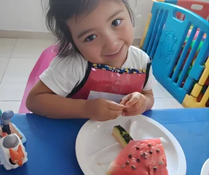 Niña sonriendo mientras come una porción de sandía en un plato blanco.
